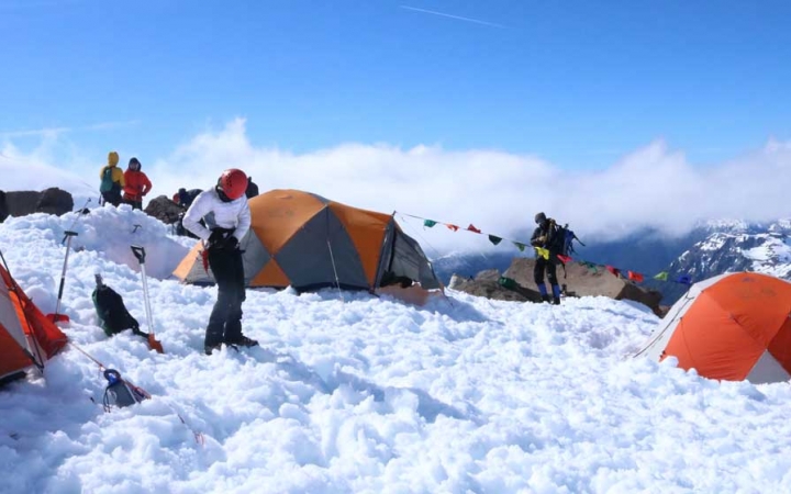 High on a mountainous landscape, tents rest in the snow of a campsite. People are standing around wearing cold-weather gear.